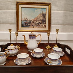 May include: A porcelain tea set featuring a white teapot, a lidded sugar bowl, a creamer, and three teacups with matching saucers. Each piece is decorated with a blue and gold rim pattern and gold handles. Four brass candlesticks stand nearby.