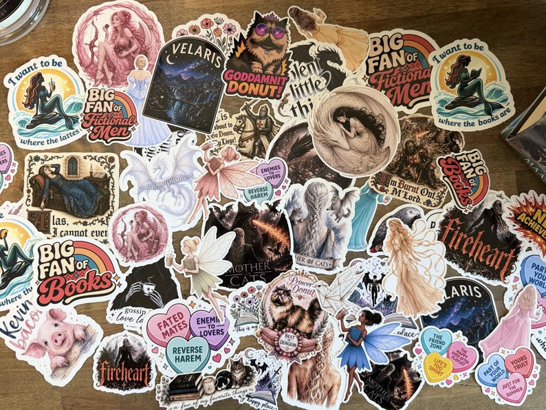 May include: A collection of assorted vinyl stickers featuring various illustrations, including fantasy characters, mythical creatures, animals, and text-based designs like Big Fan of Fictional Men, Velaris, and Fireheart, arranged in a dense, overlapping cluster.