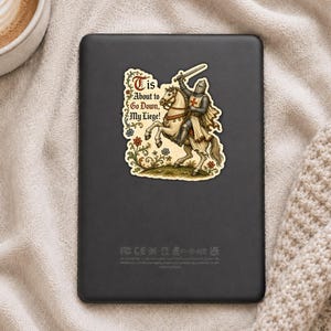 May include: A rectangular black e-reader featuring a sticker of a medieval knight in armor riding a white horse, holding a sword aloft. The sticker includes the text 'Tis About to Go Down, My Liege!' surrounded by colorful floral illustrations.