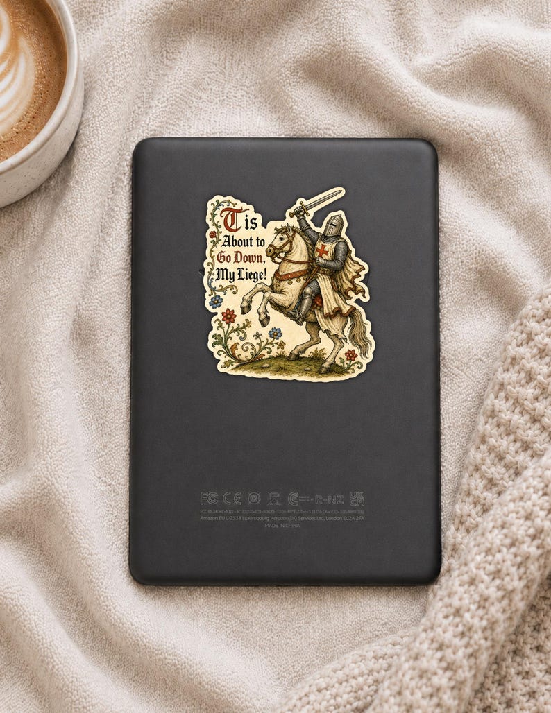 May include: A rectangular black e-reader featuring a sticker of a medieval knight in armor riding a white horse, holding a sword aloft. The sticker includes the text 'Tis About to Go Down, My Liege!' surrounded by colorful floral illustrations.