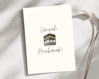 May include: A white greeting card featuring the text Umrah Mubarak in a dark grey script font. Centered between the two lines of text is a small, detailed illustration of the Kaaba, depicted in black and gold tones with a square structure.