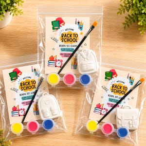 May include: Three clear plastic bags containing a Back to School activity kit. Each kit includes a printed card with school graphics, a white backpack-shaped plaster figure, a black paintbrush, and three small paint pots in yellow, pink, and blue.