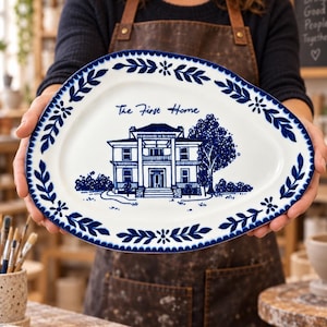 May include: An irregular oval-shaped white ceramic platter featuring a blue hand-painted illustration of a house with a large tree. The text "The First Home" is written above the house, and the rim is decorated with a blue leaf and floral pattern.