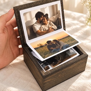 May include: A dark brown wooden box containing a stack of accordion-style photographs. The top photo shows a couple sitting on a couch, while the second photo displays the same couple sitting in a grassy field during sunset.