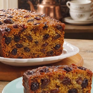 May include: A rectangular loaf of fruit cake with a golden-brown crumb and numerous dark raisins throughout. A single slice of the same fruit cake sits on a light blue plate in the foreground, showing a dense texture with visible dried fruit pieces.