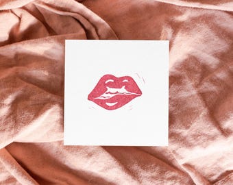 May include: A square white card featuring a centered, textured pink graphic of lips. The lips are depicted with a slight smile, showing a hint of teeth, and are rendered in a block-print style with visible ink texture and subtle carved lines.