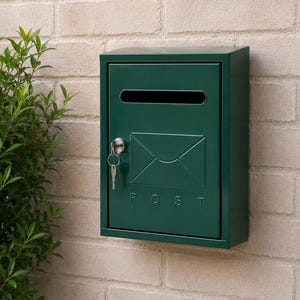 Op de afbeelding: Een donkergroene metalen wandbrievenbus met een horizontale gleuf aan de bovenkant, een verhoogd envelopicoon in het midden en het woord POST eronder in reliëf. Een zilveren sleutel zit in het ronde slot aan de linkerkant van de deur.