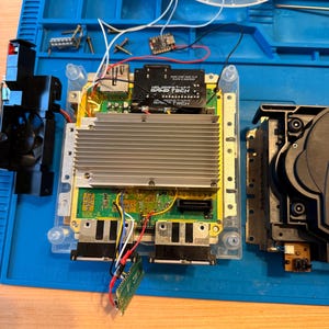 May include: An open video game console with a green circuit board, a large silver aluminum heatsink, and a black Game Tech HDMI adapter board. The internal components are exposed, featuring various wires, a small fan, and a green microcontroller board.