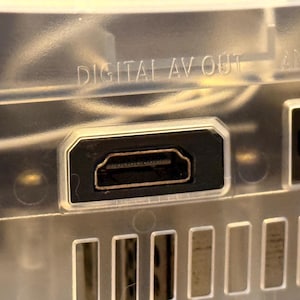 May include: A clear plastic electronic device housing featuring a black HDMI port centered below the embossed text DIGITAL AV OUT. The port is rectangular with beveled corners, and a series of vertical ventilation slots are visible below it.