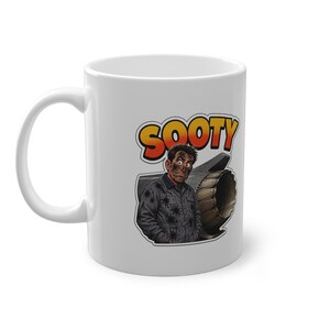 May include: A white ceramic mug featuring a graphic illustration of a person covered in soot, wearing a dark jumpsuit, standing in front of a jet engine. Above the figure, the word SOOTY is printed in bold, orange and yellow gradient lettering.