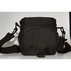 May include: A black camera bag with a structured rectangular shape, featuring a top zippered closure, a front pocket, side mesh compartments, and an adjustable shoulder strap attached with black plastic clips and hardware on both sides.