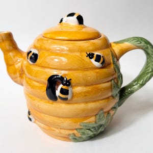 May include: A ceramic teapot shaped like a yellow beehive with a matching lid. The teapot features raised black and yellow bee figures on the exterior, a green handle textured like leaves, and a spout extending from the side of the hive body.