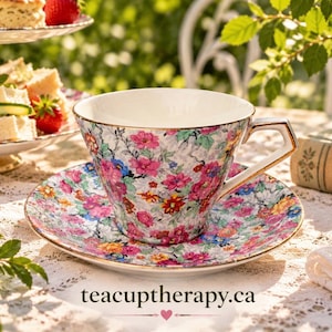 May include: A porcelain teacup and saucer set featuring a vibrant floral pattern with pink, red, orange, and blue flowers on a white background, accented with gold trim on the rim and handle. The text teacuptherapy.ca is printed at the bottom.