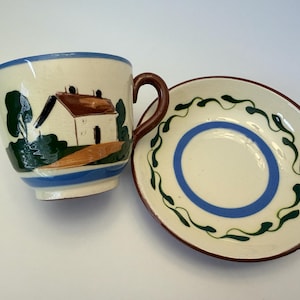 Pode incluir: Um conjunto de chávena e pires em cerâmica com uma base de cor creme pintada à mão. A chávena apresenta uma casa com telhado castanho, árvores verdes e um rebordo azul. O pires é decorado com uma banda circular azul e um padrão de videira.