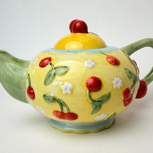 Peut inclure: Une théière en céramique avec un corps jaune orné d'un motif en relief de cerises et de fleurs blanches. Elle possède une anse et un bec verseur vert clair, une base et un bord bleu clair, et un couvercle jaune surmonté d'un petit bouton rouge.