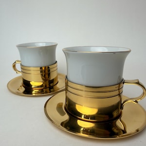 May include: Two white porcelain cups with matching saucers, each featuring a wide, polished gold-tone metal base and handle. The gold metal band wraps around the lower portion of the white cup, while the saucer is entirely finished in gold.