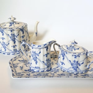 May include: A white porcelain tea set featuring a blue floral pattern with a lattice design. The set includes a teapot with a lid, a creamer jug, a lidded sugar bowl, and a rectangular serving tray, all with a vertical ribbed texture.