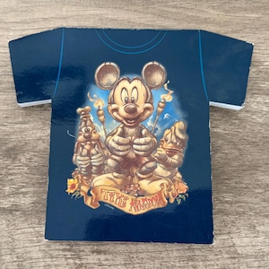 May include: A dark blue t-shirt shaped card featuring a graphic of Mickey Mouse, Goofy, and Donald Duck depicted as wooden tiki carvings. The characters are surrounded by flaming torches and tropical flowers, with the text Tiki Kingdom below them.