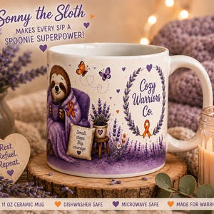 May include: A white ceramic mug featuring a sloth illustration, lavender sprigs, and the text 'Cozy Warriors Co.' with an orange ribbon. The design includes a small sign reading 'Small steps. Big courage.' and purple hearts. The mug is 11 oz.