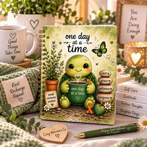 May include: A spiral-bound notebook featuring a green turtle illustration holding a mug with the text 'one day at a time'. The cover includes a potted plant, a stack of stones labeled 'rest', 'grace', 'patience', 'progress', and a butterfly.