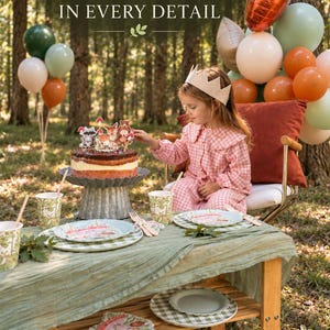 May include: A woodland party table set for 8 guests featuring a cake topper with forest animals, patterned paper plates, cups, and napkins. The set includes a sage green table runner and is displayed on a wooden table with balloons in the background.