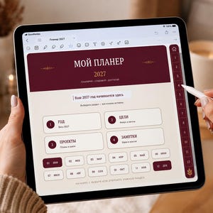 Può includere: Un tablet digitale che mostra un'interfaccia di pianificazione 2027 con un'intestazione rosso scuro e schede di navigazione. Lo schermo presenta testo in russo tra cui 'МОЙ ПЛАНЕР', 'ГОД', 'ЦЕЛИ', 'ПРОЕКТЫ' e 'ЗАМЕТКИ', oltre a una griglia mensile da 01 a 12.