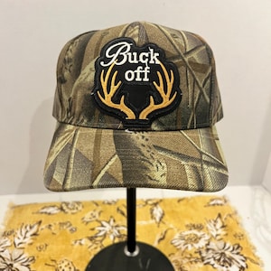 May include: A camouflage baseball cap featuring a black patch on the front. The patch displays the white text Buck off above a pair of embroidered golden-brown deer antlers. The cap has a structured crown and a curved brim with a woodland pattern.