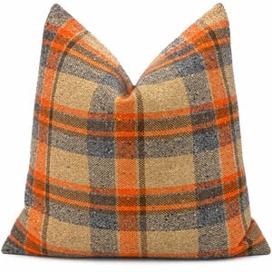 May include: A square scatter cushion featuring a woven plaid pattern in shades of bright orange, charcoal grey, and tan. The fabric has a visible textured weave with small flecks of darker colour throughout the material, creating a multi-tonal appearance.