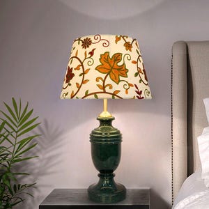 May include: A table lamp with a dark green, urn-shaped base and a tapered, off-white fabric lampshade. The shade features an embroidered pattern of colorful autumn leaves and swirling vines in shades of orange, brown, and green.