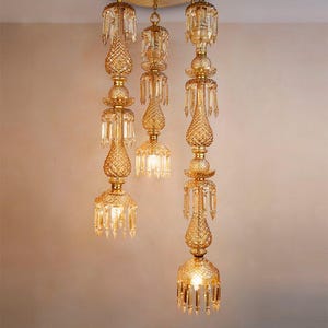 May include: A multi-pendant chandelier featuring three vertical glass fixtures suspended from a circular brass ceiling mount. Each fixture consists of amber-colored, textured glass segments with hanging crystal prisms and internal light bulbs.