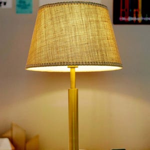 May include: A table lamp with a cylindrical brass-colored metal base and a circular stand. The lamp features a tapered, textured tan fabric lampshade with a decorative stitched trim along the top and bottom edges, emitting a warm light.
