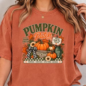 Peut inclure: Un t-shirt couleur rouille avec un motif de citrouilles dans une caisse en bois et un nœud à carreaux noir et blanc. Le texte indique PUMPKIN Patch, FARM FRESH, FRESH FROM THE PATCH, AUTUMN HARVEST GOOD VIBES, et PUMPKINS CORN MAZE APPLE CIDER HAYRIDES.