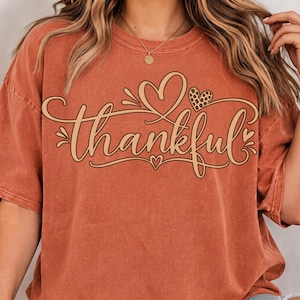 Peut inclure: Un t-shirt à manches courtes de couleur rouille avec le mot thankful écrit dans une police cursive. Le motif comprend un grand contour de cœur et un plus petit cœur à motif léopard. Le t-shirt présente un col rond et un aspect délavé.