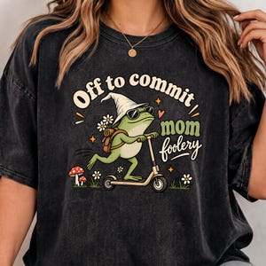 Puede incluir: Una camiseta gris carbón con un gráfico de una rana verde con sombrero puntiagudo blanco y gafas de sol montada en un patinete. El texto dice Off to commit mom foolery, junto a ilustraciones de margaritas blancas y setas rojas.