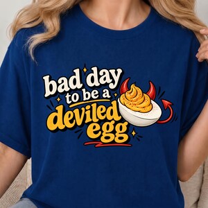 Peut inclure: Un t-shirt bleu marine avec une impression graphique représentant un œuf farci doté de cornes et d'une queue de diable rouges. Le texte "bad day to be a deviled egg" est écrit dans une police jaune et blanche stylisée avec des étoiles.