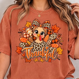 Peut inclure: Un t-shirt couleur rouille avec un motif de dinde portant un nœud à imprimé léopard et des plumes aux motifs variés, incluant du tartan, des pois et des fleurs. Le mot Thankful est imprimé en lettres cursives orange sous la dinde.