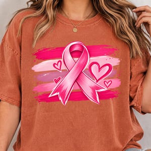 Peut inclure: Un t-shirt à manches courtes de couleur rouille avec une impression graphique d'un ruban de sensibilisation rose centré sur des coups de pinceau horizontaux dans des tons de rose. Le motif inclut trois cœurs roses de tailles différentes.