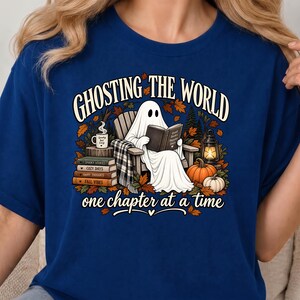 Peut inclure: Un t-shirt bleu foncé avec un motif représentant un fantôme blanc assis dans une chaise en bois en train de lire un livre. Le texte indique GHOSTING THE WORLD one chapter at a time. L'image inclut des livres, une tasse, une lanterne et des citrouilles.