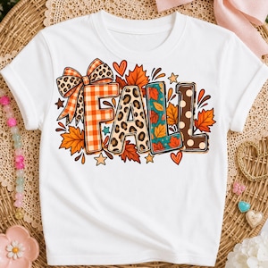 Peut inclure: Un t-shirt blanc à manches courtes avec une impression graphique du mot FALL en grandes lettres. Les lettres sont décorées de motifs à carreaux orange, léopard, feuilles orange sur fond bleu sarcelle et pois marron, avec un nœud léopard.