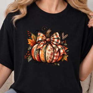 Peut inclure: Un t-shirt noir avec un motif de citrouille ornée d'un nœud à carreaux. La citrouille présente des segments orange, verts et à motifs de feuilles. Elle est entourée de feuilles d'automne, de baies et d'un petit cœur à motif léopard.
