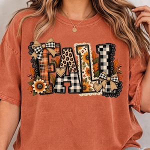 Peut inclure: Un t-shirt couleur rouille avec le mot FALL en grandes lettres majuscules. Chaque lettre présente un motif différent, notamment du tartan orange, des taches de léopard, des tournesols et un vichy noir et blanc, avec deux nœuds.
