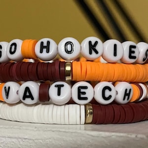 Pode incluir: Uma pilha de quatro pulseiras de contas com discos de argila planos nas cores bordô, laranja e branco. Duas pulseiras incluem contas esféricas brancas com letras pretas que formam GO HOKIES e VA TECH, acentuadas por pequenos espaçadores de metal dourado.