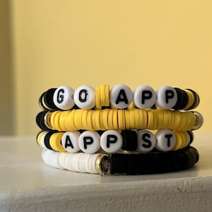 Puede incluir: Una pila de cuatro pulseras hechas de cuentas de arcilla planas y circulares en negro, amarillo y blanco. La pulsera superior tiene cuentas de letras blancas que deletrean GO APP, y la tercera tiene cuentas de letras que deletrean APP ST.