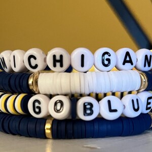 Puede incluir: Una pila de cuatro pulseras de cuentas con discos de arcilla en azul marino, blanco y amarillo con espaciadores dorados. Dos pulseras incluyen cuentas redondas blancas con letras negras que forman las palabras MICHIGAN y GO BLUE.