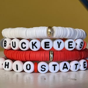 May include: A stack of three beaded bracelets featuring white and red flat circular clay beads. The middle bracelet displays white spherical beads with black letters spelling BUCKEYES, while the bottom bracelet spells OHIO STATE.