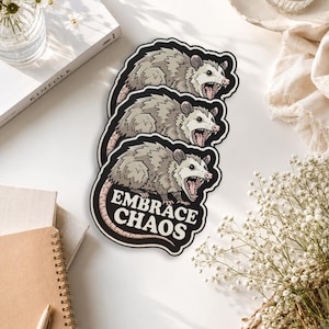 May include: A die-cut sticker featuring three stacked illustrations of a grey and white opossum with an open mouth showing teeth. The bottom opossum is positioned above bold white text that reads EMBRACE CHAOS on a black background.