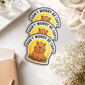 May include: Three circular stickers featuring a brown capybara illustration centered against a yellow sun background. The text DON'T WORRY BE CAPY is printed in black capital letters along the top edge of each sticker, with green leaves at the base.