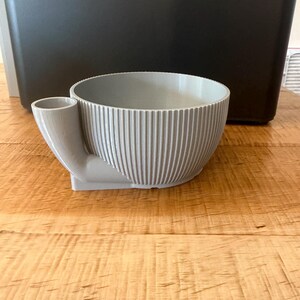 May include: A light gray, circular bowl with a ribbed, vertical texture on the exterior. The bowl features a small, cylindrical attachment on one side that extends outward and upward, creating a distinct, asymmetrical profile for this container.
