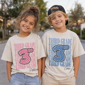 May include: Two beige short-sleeve t-shirts featuring the repeating text THIRD GRADE in pink or blue. Each shirt displays a large checkered number 3 with the letters RD. The pink shirt includes the name Sophia, and the blue shirt includes Liam.