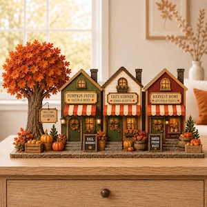 Puede incluir: Una escena decorativa de pueblo en miniatura con tres tiendas con toldos de rayas naranjas y blancas, ventanas iluminadas y letreros de otoño. Un gran árbol con hojas naranjas se encuentra a la izquierda, acompañado de calabazas.